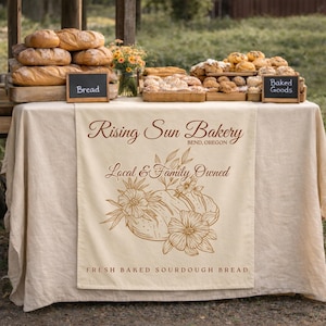 May include: A display of baked goods at a bakery, including loaves of bread and pastries. The table is covered with a cream-coloured tablecloth and a banner with the text "Rising Sun Bakery, Bend, Oregon, Local & Family Owned, Fresh Baked Sourdough Bread."
