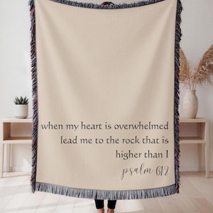 May include: A beige throw blanket with a dark blue and red fringe. The blanket has the text "when my heart is overwhelmed lead me to the rock that is higher than I psalm 61:2" in black script.