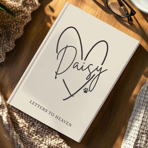 Könnte beinhalten: Ein cremefarbenes Notizbuch mit dem Namen "Daisy" in Herzform. Es zeigt auch einen Pfotenabdruck und den Text "Letters to Heaven". Das Notizbuch liegt auf einer Holzoberfläche, mit einer Brille im Hintergrund.