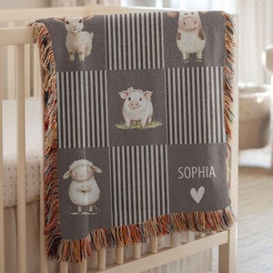 May include: A grey and white woven baby blanket with a farm animal theme. The blanket features a lamb, cow, pig, and sheep, with vertical stripes and a personalised name "SOPHIA" and a heart symbol. The blanket has a fringed edge.