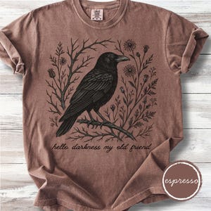 May include: Espresso-colored t-shirt featuring a black raven perched on a branch, surrounded by floral and branch illustrations. The phrase "hello darkness my old friend" is printed below the raven. The shirt has a crew neck and short sleeves.