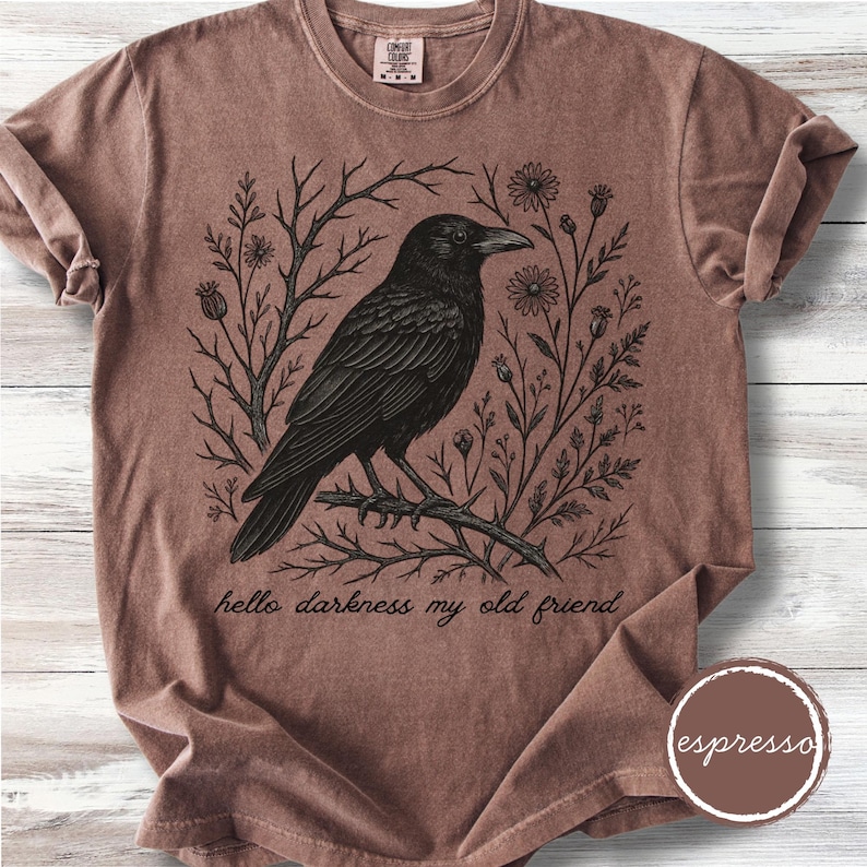 May include: Espresso-colored t-shirt featuring a black raven perched on a branch, surrounded by floral and branch illustrations. The phrase "hello darkness my old friend" is printed below the raven. The shirt has a crew neck and short sleeves.