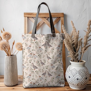 May include: A beige tote bag with a repeating pattern of gray birds wearing bow ties, interspersed with green leaves and red berries. The bag has brown leather handles and is displayed on a wooden stand.