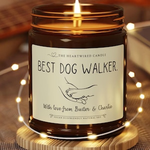 May include: A lit brown glass jar candle on a wooden coaster. The candle has a white label with the text "BEST DOG WALKER" and a line drawing of a hand holding a paw. The text "With love from Buster & Charlie" is also on the label.