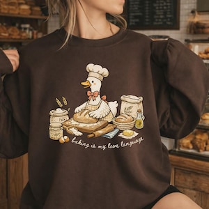 May include: A brown sweatshirt featuring a cartoon goose chef baking bread. The goose wears a chef's hat and apron, with a pink bow tie. The text "baking is my love language" is below the goose. Other baking supplies are also shown.