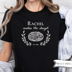 May include: Black t-shirt with white text that reads "Rachel makes the dough" and a stylised image of a seashell with the text "Est. 2024" below it.
