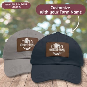 May include: Two baseball caps with a brown leather patch featuring a tractor and the text "Marigold Farms". One cap is grey and the other is navy blue.