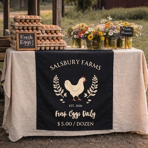 May include: A farm stall display with fresh eggs and flowers. The table has a black runner with "Salsbury Farms" and a hen graphic. A chalkboard sign reads "Fresh Eggs." Jars of colourful flowers are arranged on the table.