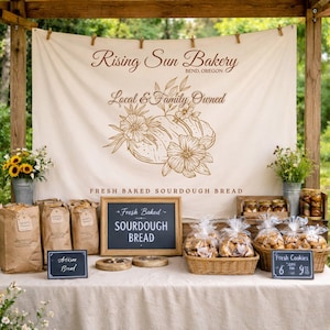 Puede incluir: Un puesto de panadería con una pancarta que dice "Rising Sun Bakery, Bend, Oregon, Local & Family Owned." Se exhiben cestas de galletas y hogazas de pan. Un letrero de pizarra anuncia pan de masa madre recién horneado. Flores en cubos de metal.
