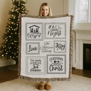 May include: A beige throw blanket with a fringed edge, featuring black text and illustrations in postage stamp-like frames. The text includes phrases like "Blessed," "Oh Holy Night," "Love," "Joy," and "Christmas Begins With Christ."