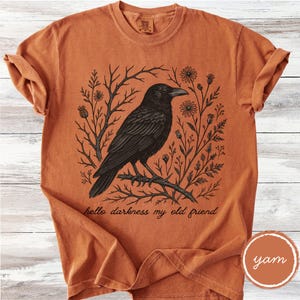 May include: Burnt orange t-shirt featuring a black crow perched on a branch, surrounded by floral and branch illustrations. The text "hello darkness my old friend" is printed below the crow. The shirt has rolled-up sleeves.