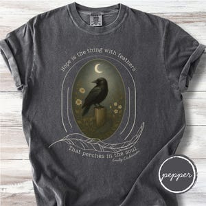 K&ouml;nnte beinhalten: Dunkelgraues T-Shirt mit einem ovalen Design, das eine schwarze Kr&auml;he auf einem Holzpfosten unter einem Halbmond zeigt. Der Text "Hope is the thing with feathers" und "That perches in the soul" ist auf dem Shirt aufgedruckt, zusammen mit dem Namen "Emily Dickinson".