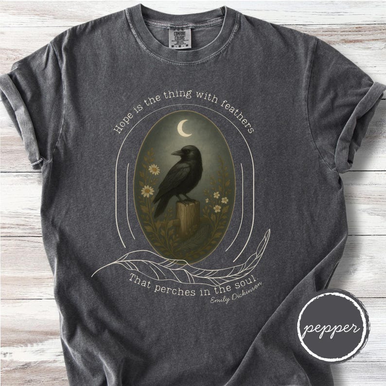K&ouml;nnte beinhalten: Dunkelgraues T-Shirt mit einem ovalen Design, das eine schwarze Kr&auml;he auf einem Holzpfosten unter einem Halbmond zeigt. Der Text "Hope is the thing with feathers" und "That perches in the soul" ist auf dem Shirt aufgedruckt, zusammen mit dem Namen "Emily Dickinson".