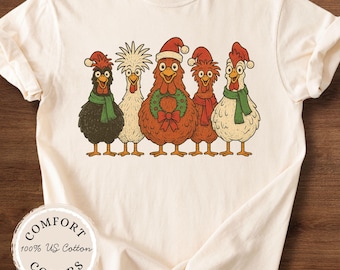 May include: Cream-colored t-shirt featuring a graphic of five cartoon chickens wearing Santa hats and scarves. The central chicken has a Christmas wreath. The shirt has the text "Comfort Colors" and "100% US Cotton".