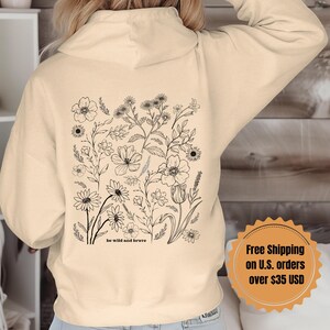 May include: A cream-colored hoodie with a black floral design on the back. The design includes a variety of flowers and leaves, and the text "be wild and brave".
