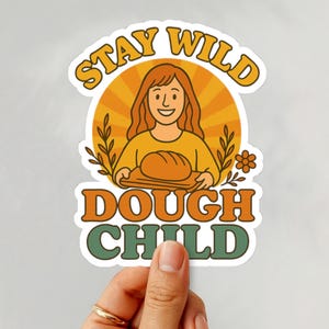 May include: A die-cut sticker featuring a person with long hair holding a loaf of bread on a wooden board. The background is a yellow sunburst with wheat stalks and a flower. The text reads STAY WILD DOUGH CHILD in orange and green.