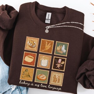 May include: A dark brown sweatshirt with a nine-square graphic of baking-related images, including bread and utensils. The phrase "baking is my love language" is printed at the bottom. A silver necklace with a sun-shaped pendant is draped across the sweatshirt.