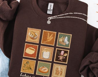 May include: A dark brown sweatshirt featuring a nine-square graphic of baking-related images, including bread and utensils. The phrase "baking is my love language" is printed at the bottom. A silver necklace with a sun-shaped pendant is draped across the sweatshirt.