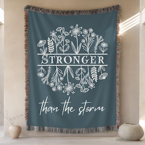 May include: A teal throw blanket with a floral design and the words "STRONGER than the storm" in white script. The blanket has a fringed edge with brown and tan threads. The design features various flowers and leaves.