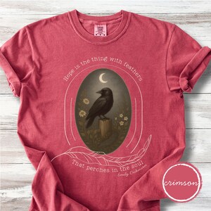 K&ouml;nnte beinhalten: Karmesinrotes T-Shirt mit einem Aufdruck eines schwarzen Raben auf einem Holzpfosten, umrahmt von einem Oval. Das Design beinhaltet einen Halbmond, Blumen und das Zitat "Hope is the thing with feathers that perches in the soul."