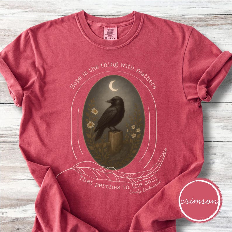 K&ouml;nnte beinhalten: Karmesinrotes T-Shirt mit einem Aufdruck eines schwarzen Raben auf einem Holzpfosten, umrahmt von einem Oval. Das Design beinhaltet einen Halbmond, Blumen und das Zitat "Hope is the thing with feathers that perches in the soul."