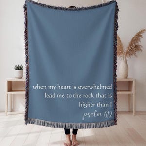 Puede incluir: Una manta tejida azul con un borde con flecos. La manta presenta texto blanco que dice "when my heart is overwhelmed lead me to the rock that is higher than I psalm 612."