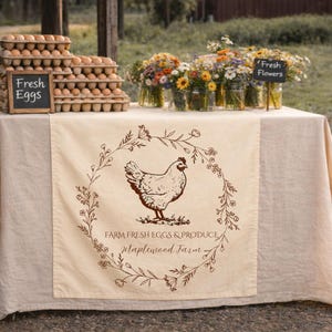 May include: A farm stand display with fresh eggs, flowers, and a cream-colored table runner. The runner features a brown hen illustration within a floral wreath, with text that reads "Farm Fresh Eggs & Produce, Maplewood Farm."