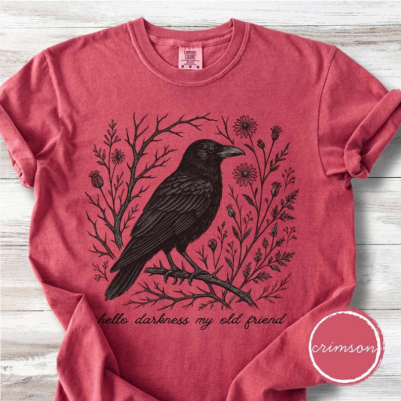 May include: Crimson-colored t-shirt featuring a black illustration of a crow perched on a branch, surrounded by floral and botanical elements. The text "hello darkness my old friend" is printed below the crow.