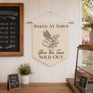 May include: A cream-coloured banner with the text "BAKED AT DAWN" and "Gone Too Soon SOLD OUT" in black script. The banner features a black illustration of bread, wheat, and flowers. A chalkboard menu and a small sign that says "Sorry! We're SOLD OUT" are also visible.