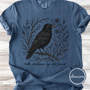 May include: A denim blue t-shirt featuring a detailed black illustration of a raven perched on a branch, surrounded by floral and botanical elements. The text "hello darkness my old friend" is printed below the raven.