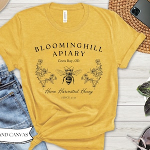 Könnte beinhalten: Senfgelbes T-Shirt mit einer schwarzen Grafik einer Biene, Blumen und dem Text "Bloominghill Apiary, Coos Bay, OR, Home Harvested Honey, Since 2021." Das Shirt ist aus weichem Material.