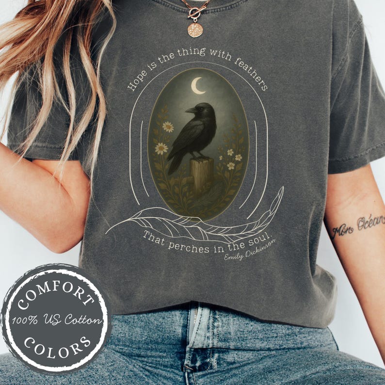 K&ouml;nnte beinhalten: Dunkelgraues T-Shirt mit einem Raben-Motiv, umgeben von Blumen und Mond. Der Text lautet "Hope is the thing with feathers" und ein Zitat von Emily Dickinson. Hergestellt aus 100% US-Baumwolle.