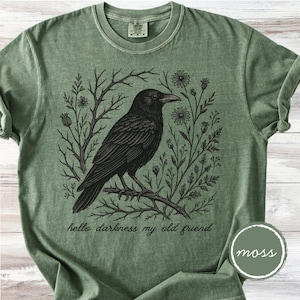 May include: A moss green t-shirt featuring a detailed black illustration of a raven perched on a branch, surrounded by floral and botanical elements. The text "hello darkness my old friend" is below the raven. A circular "moss" label is in the lower right corner.