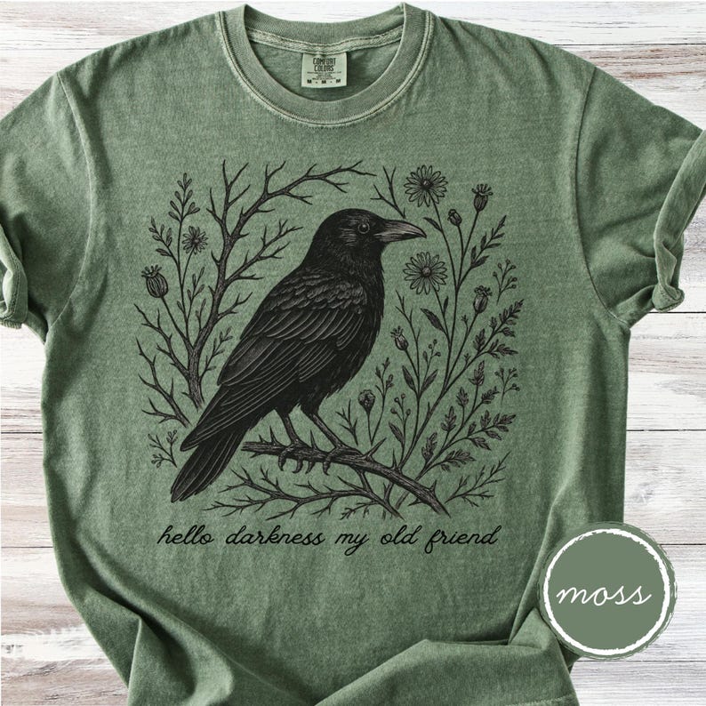 May include: A moss green t-shirt featuring a detailed black illustration of a raven perched on a branch, surrounded by floral and botanical elements. The text "hello darkness my old friend" is below the raven. A circular "moss" label is in the lower right corner.