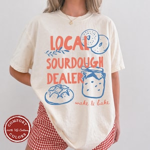 May include: Cream-colored t-shirt with the text "LOCAL SOURDOUGH DEALER" in coral and blue. The design includes illustrations of bagels, a jar of jam, and a loaf of bread. The shirt also has the words "wake & bake" in a script font.