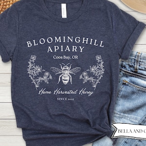 May include: Navy blue t-shirt with the text "BLOOMINGHILL APIARY Coos Bay, OR" and a detailed bee illustration. The shirt also says "Home Harvested Honey SINCE 2021". The shirt is made of soft material.