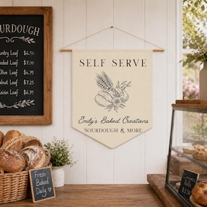 May include: A cream-colored banner with the words "SELF SERVE" and "Emily's Baked Creations" in black lettering. The banner features a bread and wheat illustration. A chalkboard menu and a basket of bread are also visible.