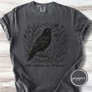 May include: Dark gray t-shirt featuring a black crow perched on a branch, surrounded by floral and branch designs. The text "hello darkness my old friend" is printed below the crow. The shirt has a crew neck and short sleeves.