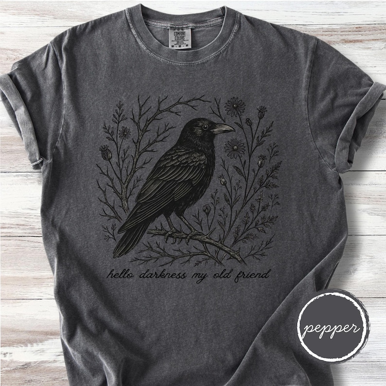 May include: Dark gray t-shirt featuring a black crow perched on a branch, surrounded by floral and branch designs. The text "hello darkness my old friend" is printed below the crow. The shirt has a crew neck and short sleeves.