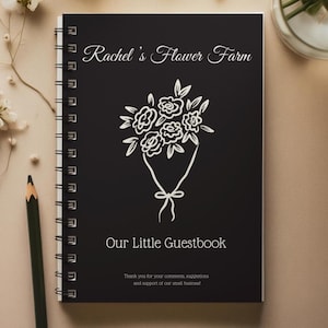 May include: A black spiral-bound guestbook with the text "Rachel's Flower Farm" and "Our Little Guestbook" in white script. A white floral bouquet illustration is centered on the cover. A pencil rests on the side.