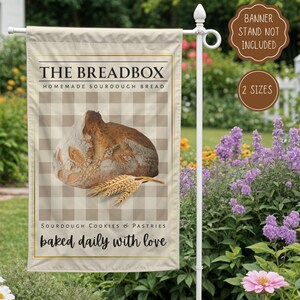 May include: A beige garden flag with the text "THE BREADBOX" and "HOMEMADE SOURDOUGH BREAD". It features a loaf of bread and wheat stalks on a checkered background. The bottom text reads "baked daily with love".
