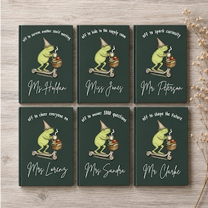 May include: Six dark green notebooks featuring a frog wearing a pointed hat, riding a scooter, and holding a mug. Each notebook displays unique text above the frog and a personalized name in white script at the bottom, including Ms. Holdan, Miss Jones, Mr. Peterson, Mrs. Lorenz, Mrs. Sandra, and Mr. Clarke.