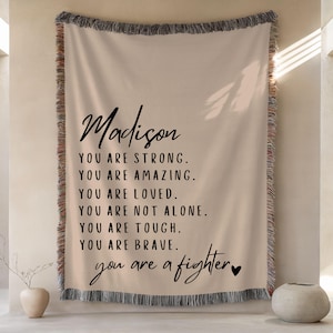May include: Beige woven throw blanket with black text. The text reads "Madison, YOU ARE STRONG. YOU ARE AMAZING. YOU ARE LOVED. YOU ARE NOT ALONE. YOU ARE TOUGH. YOU ARE BRAVE. you are a fighter". The blanket has a fringed edge.