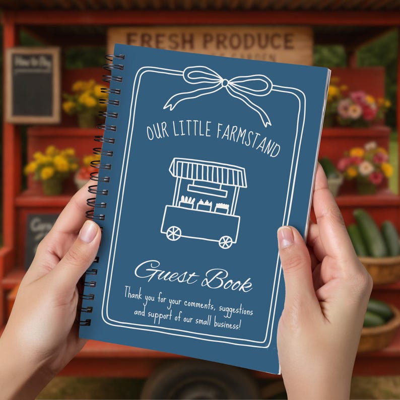 May include: A spiral-bound guest book with a dark blue cover. The cover features white text and illustrations, including "OUR LITTLE FARMSTAND" and "Guest Book." The book is held in front of a fresh produce stand.