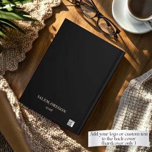 May include: A black hardcover book featuring the gold text "SALEM, OREGON" and "2026" printed on the front cover, along with a small square QR code. The book is positioned on a wooden surface next to a pair of black-rimmed eyeglasses and a white cup.