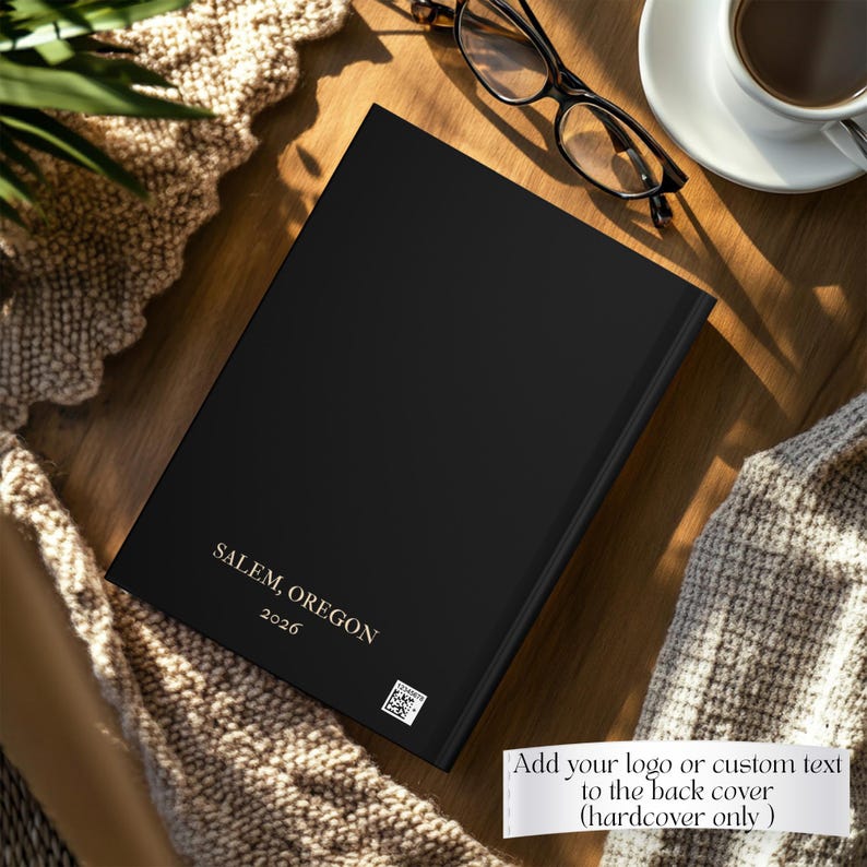 May include: A black hardcover book featuring the gold text "SALEM, OREGON" and "2026" printed on the front cover, along with a small square QR code. The book is positioned on a wooden surface next to a pair of black-rimmed eyeglasses and a white cup.