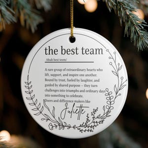 May include: A white ceramic Christmas ornament with the text "the best team" and a definition. The ornament has a gold string for hanging and is decorated with a floral design. The name "Juliette" is also included.