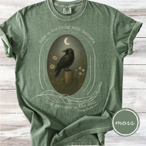 K&ouml;nnte beinhalten: Moosgr&uuml;nes T-Shirt mit einem ovalen Design, das eine schwarze Kr&auml;he auf einem Holzpfosten unter einem Halbmond zeigt. Der Text "Hope is the thing with feathers" und ein Zitat von Emily Dickinson sind ebenfalls enthalten.