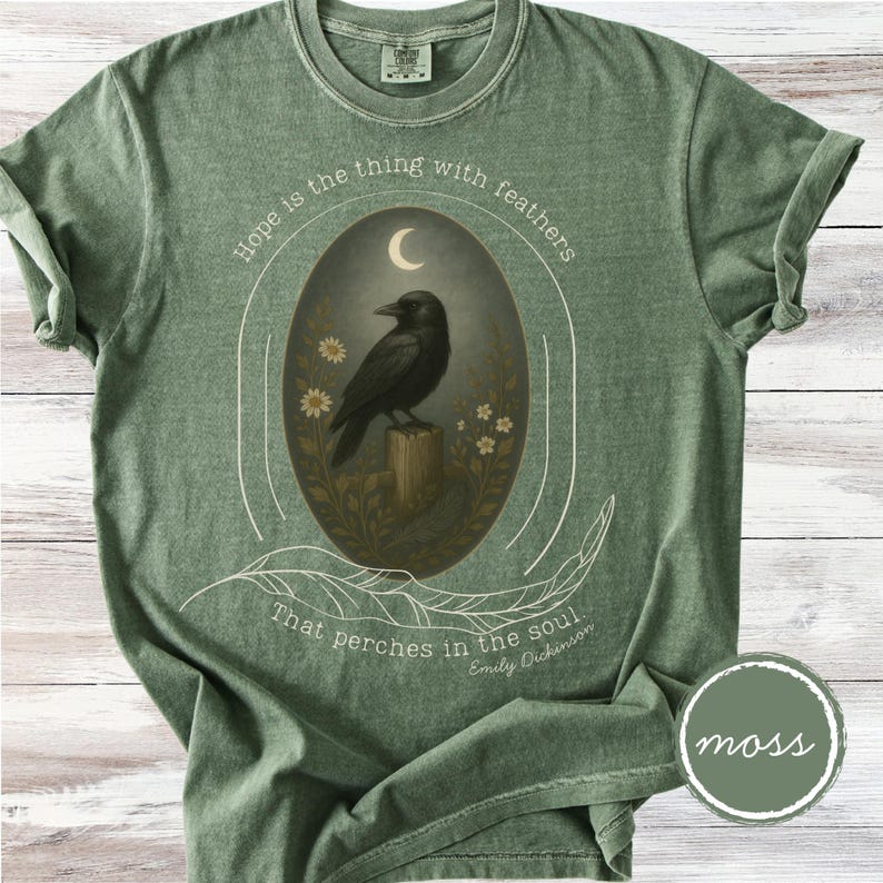 K&ouml;nnte beinhalten: Moosgr&uuml;nes T-Shirt mit einem ovalen Design, das eine schwarze Kr&auml;he auf einem Holzpfosten unter einem Halbmond zeigt. Der Text "Hope is the thing with feathers" und ein Zitat von Emily Dickinson sind ebenfalls enthalten.