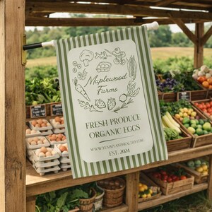 以下が含まれることがあります： 「Maplewood Farms Fresh Produce Organic Eggs」の文字と野菜や果物のイラストが描かれた、緑と白のストライプのバナー。木製の農産物スタンドの前に吊るされています。